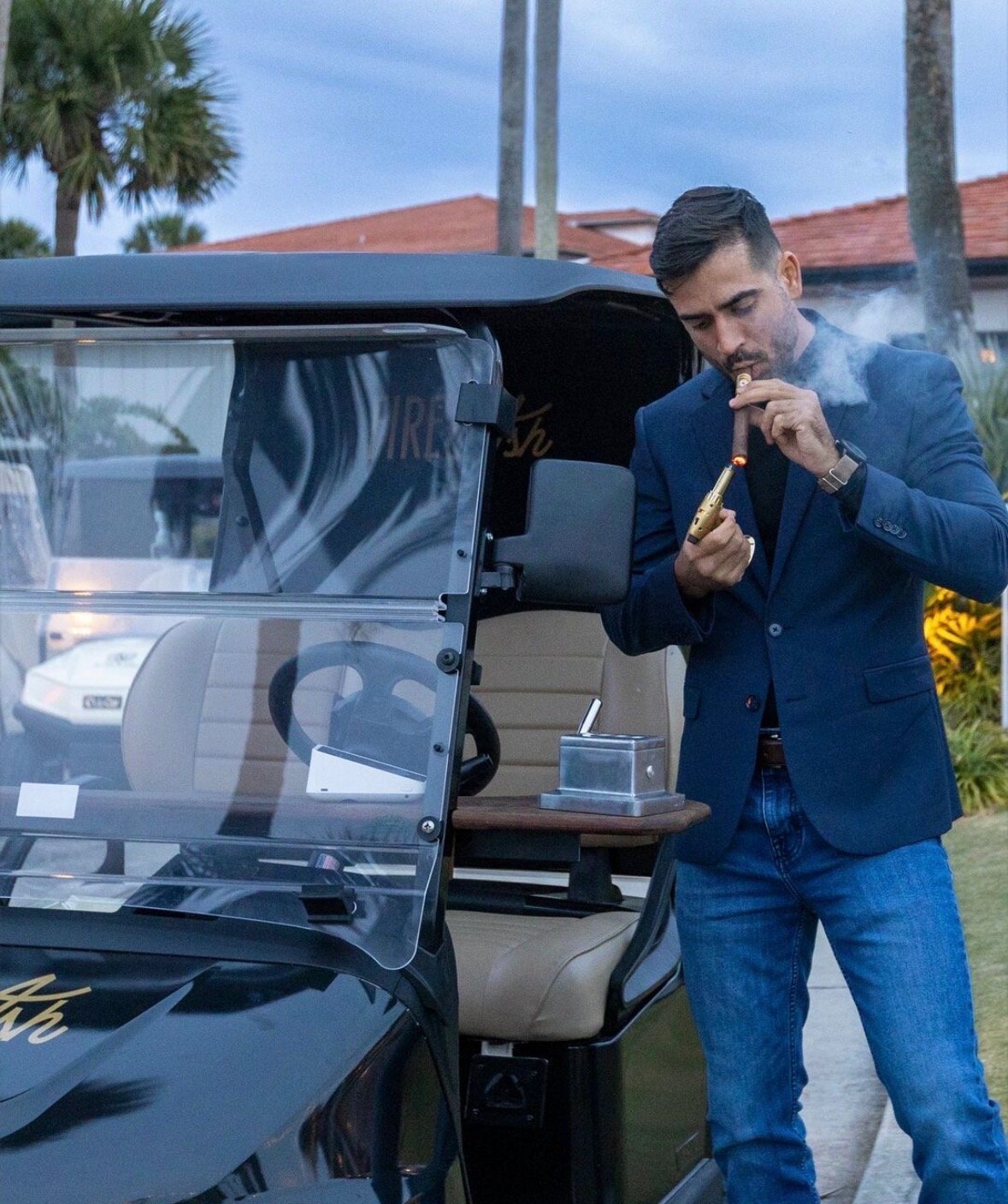 Man in blue blazer smoking cigar next to open door of black golf cart with palm trees and building in background