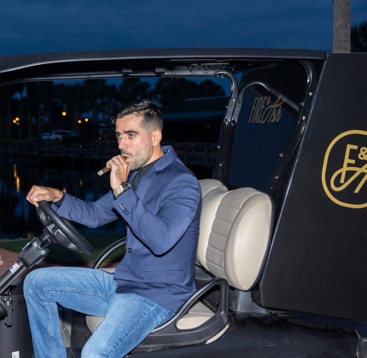 Man in blue jacket sitting in drivers seat of classic hot rod at dusk, hand on steering wheel