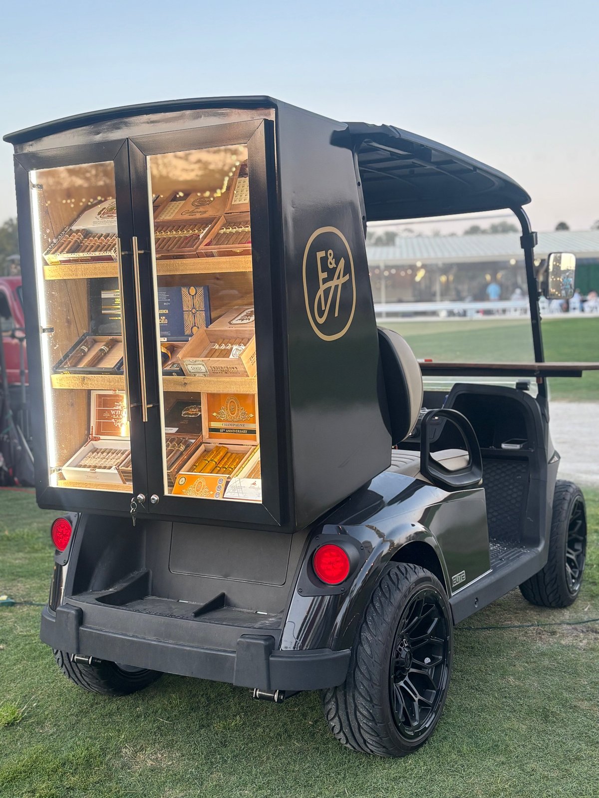 Black golf cart converted into a mobile bakery with illuminated display cases showcasing baked goods, parked on grass.